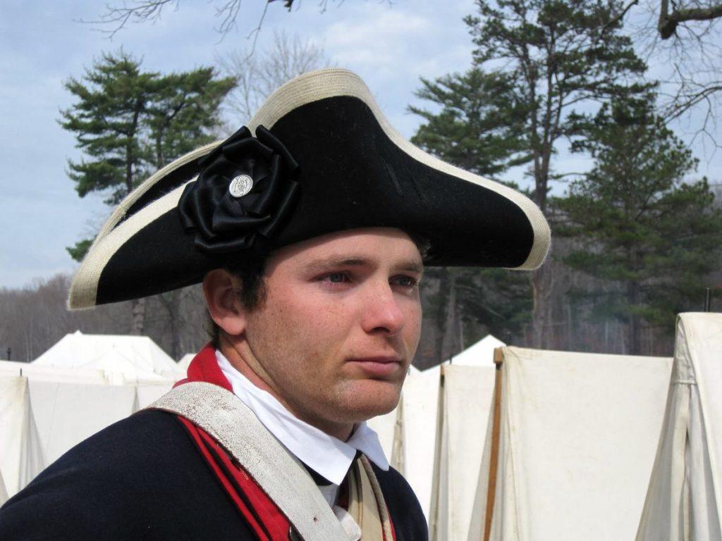 Modern color photograph of a young white man wearing a black cocked had with white trim and a rosette ribbon in black with a silver button at the center. His clothes are a white high-collared shirt, red and blue wool jacket of military design and he has a few white straps in leather and other materials going over both shoulders. In the background is a row of white military tents and further back is a tree line.